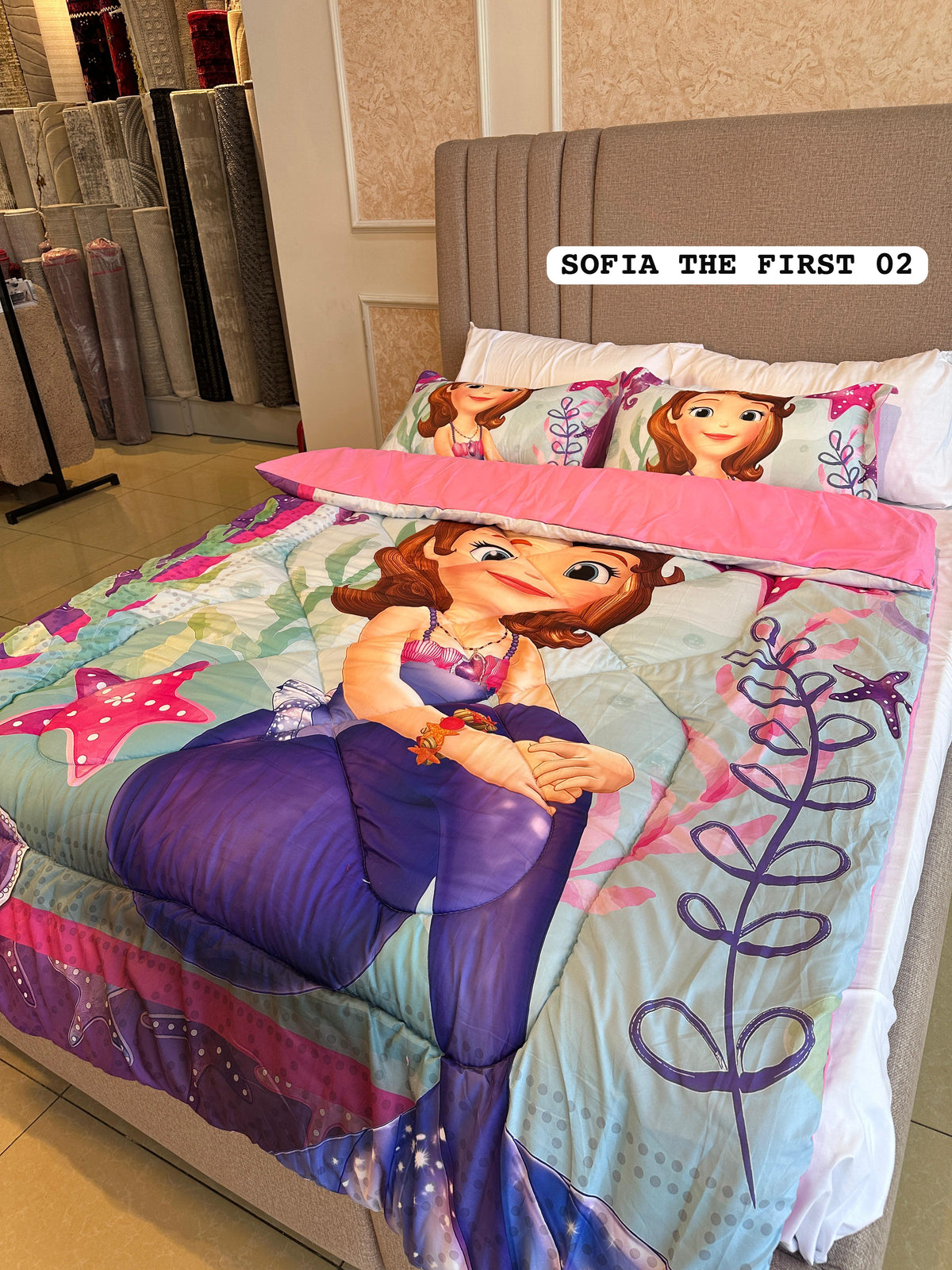 A bed made up with a Sofia the First themed bedding set, featuring a colorful mermaid print with pink accents, pillows with Sofia's face, and a pink folded backsheet. 